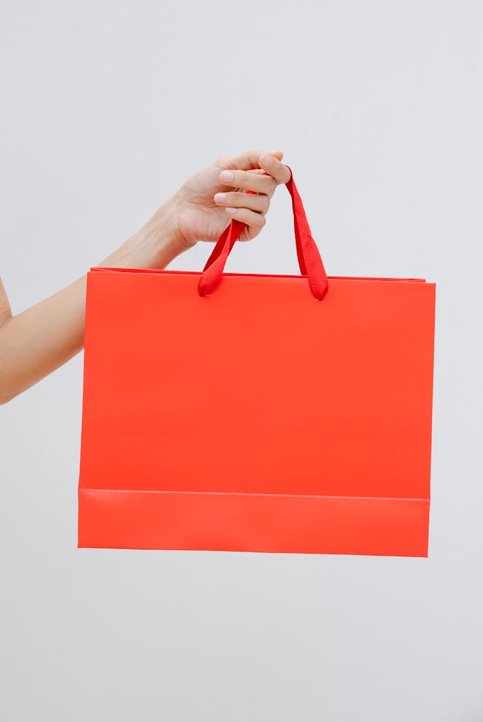 Crop unrecognizable female showing red paper shopping bag in hand against white wall